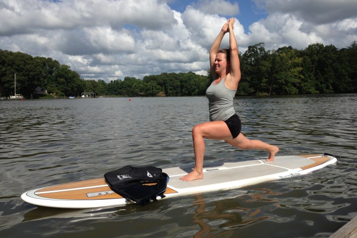 Person practicing yoga on a paddleboard in a lake with cloudy sky and trees in background.