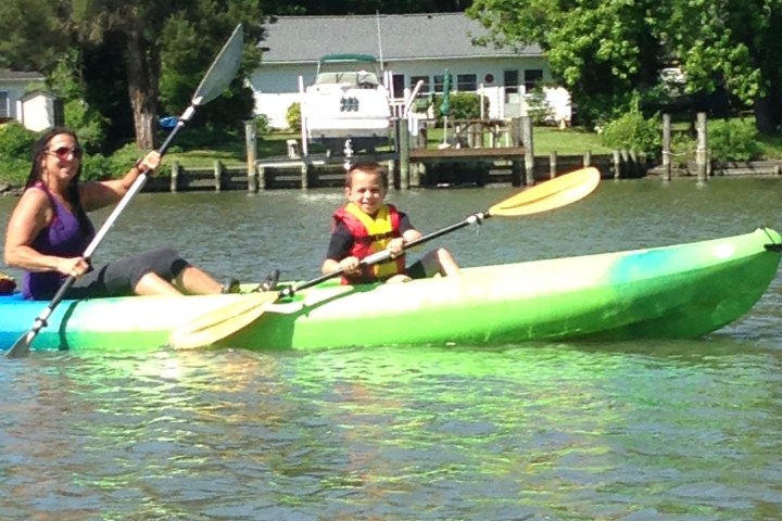 Two people kayaking on a river near houses and trees.